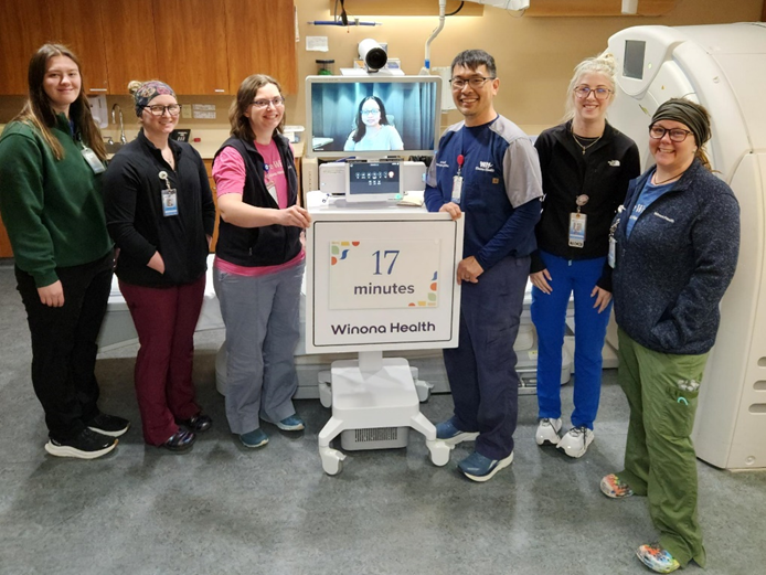 six staff members and one person on a computer screen stand in a hospital room.
