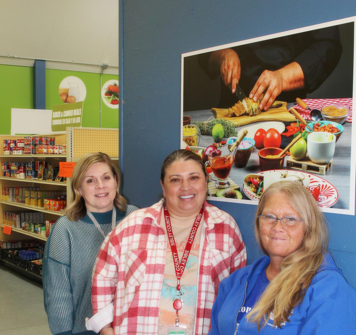 Three women stand in a food shelf with a poster of healthy foods behind them. 